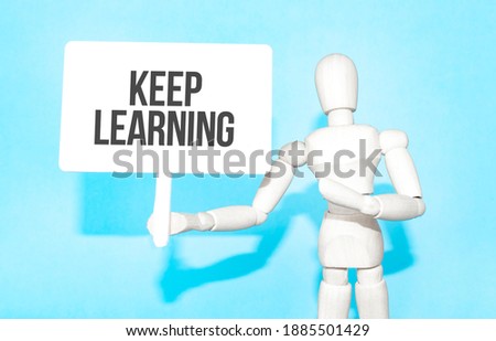 The wooden man holds a white sign with the text Keep Learning in his hands. The content of the lettering has implications for business concept and marketing.
