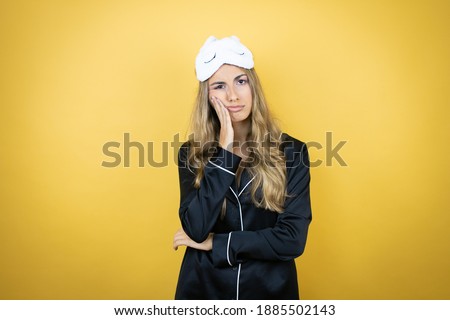 Young pretty woman wearing sleep mask and pajamas over isolated yellow background thinking looking tired and bored with crossed arms