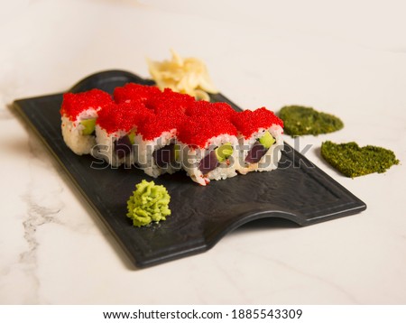 Close up of Japanese sushi roll with tuna, avocado wrapped in rice with red flying fish roe Tobiko on top. Black plate served with wasabi, ginger. Two hearts of dried herbs spices on marble background