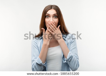 Scared caucasian girl with long chestnut hair, dressed in casual clothes, covering mouth with hands on white background. Keep silence concept.