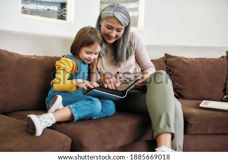 Happy grandmother showing family photo album to little smiling granddaughter while sitting on sofa at home. Generation and happy family concept