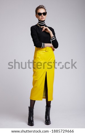 Fashion photo of a beautiful elegant young woman in a pretty yellow leather long skirt, black blouse, stylish sunglasses, accessories, boots posing over white, soft gray background. Studio Shot. 