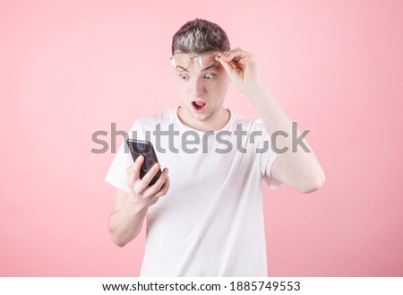Portrait of shocked young guy in white t-shirt looking at mobile phone in surprise, isolated on pink background