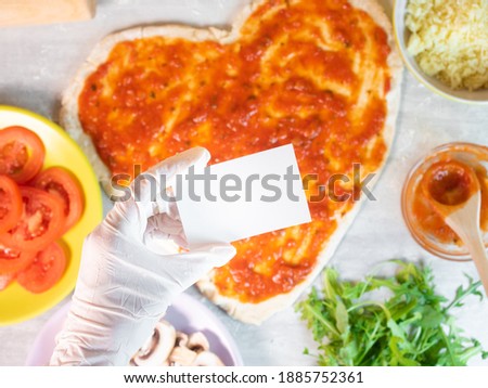 Hand in glove holding empty business card on hear shape pizza dough with tomato sauce background. Ingredients for cooking pizza with dough in heart shape