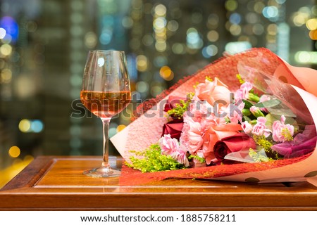 glass of red wine with bouquet of roses and carnation on wood table with blurred colorful city night lights in background. romantic concept for lovers and valentine Royalty-Free Stock Photo #1885758211