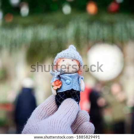 
A female hand in a mitten holds a doll with a blurry view of the Christmas tree. Making a Christmas gift doll for children. Close up view of handmade doll with accessories. High quality photo