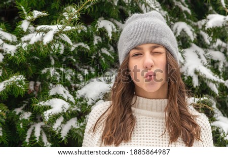 A cute girl winks and sends an air kiss on the background of a green, snowy Christmas tree. Place for an inscription. Postcard