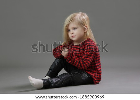 Fashion studio portrait of adorable 3 years old girl in red plaid shirt and leather black pants on grey background.