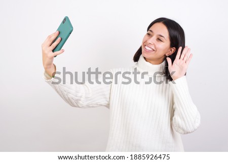 Young brunette woman wearing white knitted sweater against white background smiling and taking a selfie ready to post it on her social media.