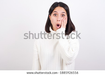 Shocked Young brunette woman wearing white knitted sweater against white background looks with great surprisment being very stunned, astonished with unexpected news, Facial expressions concept.