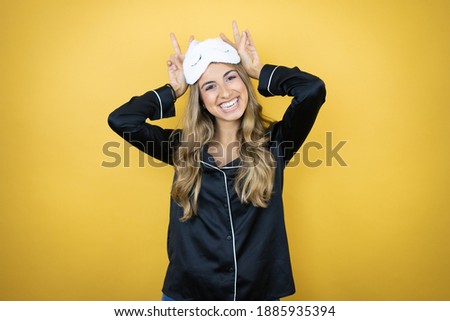 Young pretty woman wearing sleep mask and pajamas over isolated yellow background Posing funny and crazy with fingers on head as bunny ears, smiling cheerful