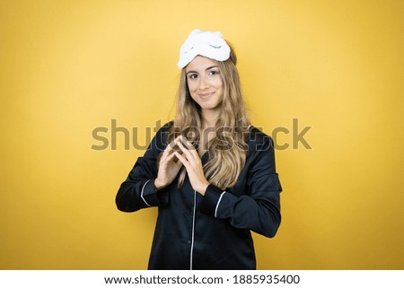Young pretty woman wearing sleep mask and pajamas over isolated yellow background with Hands together and fingers crossed smiling relaxed and cheerful. Success and optimistic