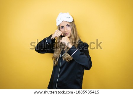 Young pretty woman wearing sleep mask and pajamas over isolated yellow background Punching fist to fight, aggressive and angry attack, threat and violence