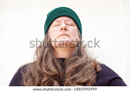 Middle aged woman wearing a navy blue sweater and a green beanie cap has long thick brown and gray hair and stands outside against a white background falling asleep                              
