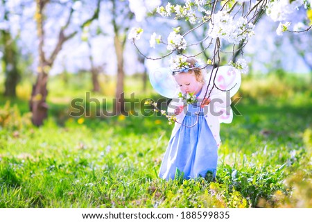 Adorable toddler girl with curly hair and flower crown wearing a magic fairy costume with a blue dress and angel wings playing in a sunny blooming fruit garden with cherry blossom and apple trees 