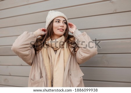 Happy beautiful young woman with smiles in a stylish outerwear with a knitted hat, fashionable jacket and a scarf stands on a wooden wall on the street. Female casual winter vintage style  Royalty-Free Stock Photo #1886099938