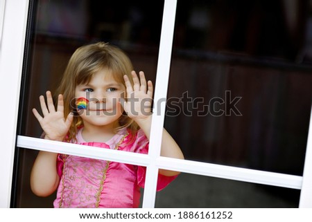 Cute lonely little toddler girl in princess dress sitting by window with rainbow with colorful colors on face during pandemic coronavirus quarantine. Children make and paint rainbows around the world