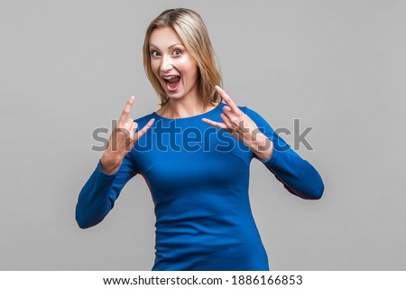 Portrait of crazy funny woman in elegant blue dress looking at camera with excited happy face and showing rock symbol with fingers up, devil horns. indoor studio shot isolated on gray background