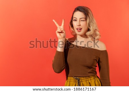 Portrait of happy young woman with blonde hair in brown blouse showing victory gesture by her fingers and playfully winking at the camera. Indoor studio shot isolated on red background