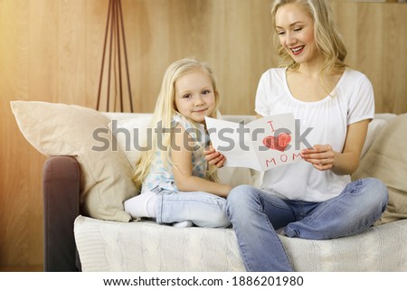 Happy mother day in sunny flat. Child daughter congratulates mom and gives her postcard with heart drawing. Family concept