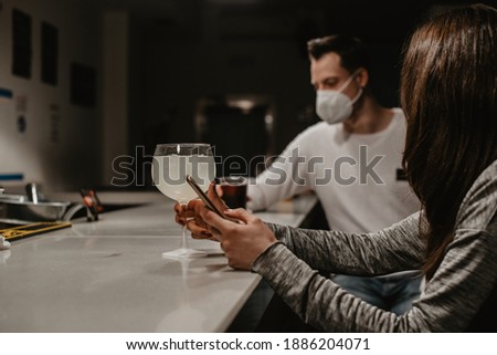 A young girl sitting at a bar counter holding her mobile phone. On the counter there is a glass of a white alcohol with lemon. At the back a young man wears a white surgical mask covering his mouth