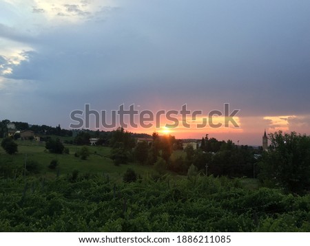 The sunset in a countryside landscape, in Italy: you can see green fields, some houses and on the right the bell tower of the church. Cloudy sky. 