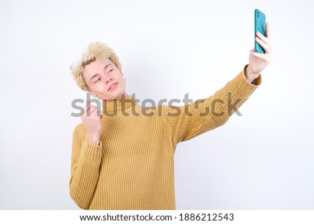 Portrait of a Young handsome Caucasian blond man standing against white background  taking a selfie to send it to friends and followers or post it on his social media.