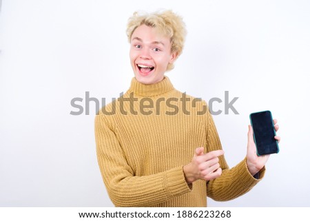 Excited Young handsome Caucasian blond man standing against white background holding and pointing with finger at smartphone with blank screen. Advertisement concept.