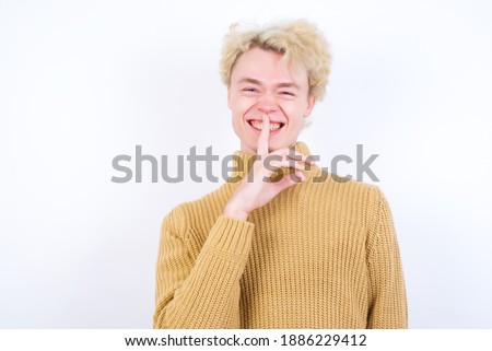 Smiling Young handsome Caucasian blond man standing against white background makes shush gesture, holds fore finger over lips hides secret. Be mute, please.