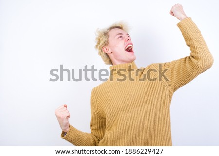 Attractive Young handsome Caucasian blond man standing against white background celebrating a victory punching the air with his fists and a beaming toothy smile