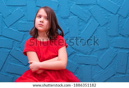 a little girl in a red dress sits against the wall and she is unhappy, on a blue background