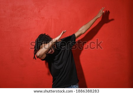 Cheerful young african american man with dreadlocks 20s in black t-shirt doing dab dance gesture hip hop hands youth sign cover hiding covering face isolated on red color background studio portrait