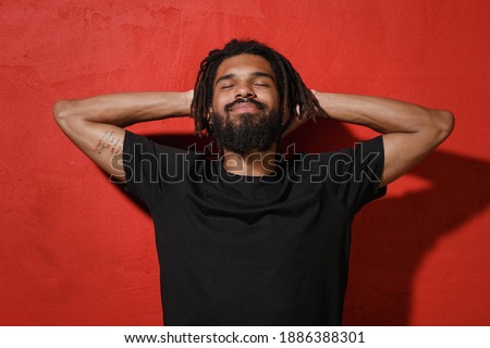 Relaxed calm smiling young african american man with dreadlocks 20s wearing black basic casual t-shirt posing sleep with hands behind head isolated on bright red color wall background studio portrait Royalty-Free Stock Photo #1886388301