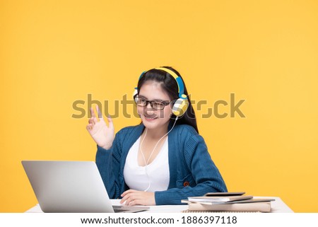 Portrait of pretty young Asian woman study wearing wireless headphones, glasses video call on laptop computer isolated over yellow background. Young girl learning online during new normal concept.