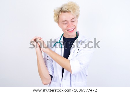 Dreamy young handsome Caucasian doctor man standing against white background with pleasant expression, closes eyes, keeps hands crossed near face, thinks about something pleasant