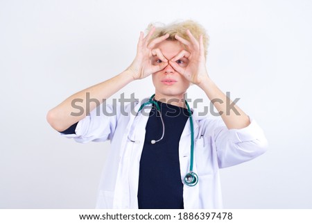 Playful excited young handsome Caucasian doctor man standing against white background showing Ok sign with both hands on eyes, pretending to wear spectacles.