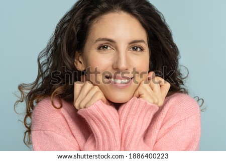 Close up of romantic young woman keep hands under chin, looking at camera, smiling, has white teeth, wear rosy sweater, has no make up, isolated on studio blue color background. Joy positive emotions