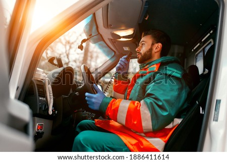 An adult handsome male paramedic is talking on a portable radio while sitting in an ambulance outside a clinic. Royalty-Free Stock Photo #1886411614