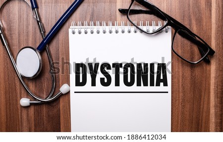 The word DYSTONIA is written on white paper on a wooden background near a stethoscope and black-framed glasses. Medical concept