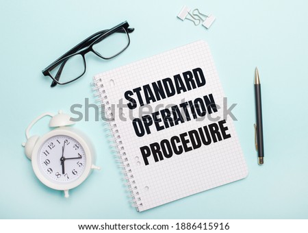 On a light blue background lie black glasses and a pen, a white alarm clock, white paper clips and a notebook with the words Standard Operating Procedure. Business concept