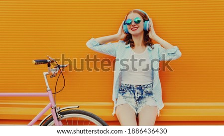 Happy young smiling woman listening to music in headphones with bicycle on an orange background