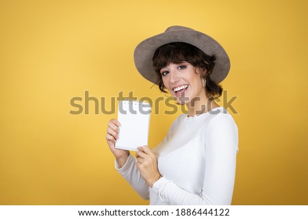Young caucasian woman wearing hat over isolated yellow background smiling and showing blank notebook in her hand