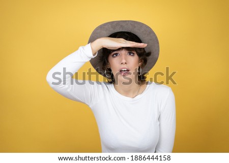 Young caucasian woman wearing hat over isolated yellow background very happy and smiling looking far away with hand over head. searching concept.