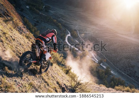 Extreme Rider climbing sand mountain top on off-road cross enduro motorcycle. Beautiful mountains landscape down on background, colourful autumn forest and river in sunshine 