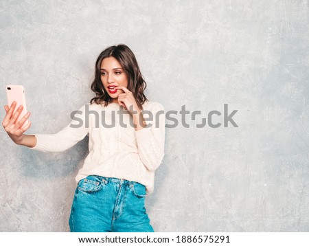 Young beautiful woman taking selfie photos. Trendy smiling model in casual summer jeans clothes. Positive female with red lips taking selfie photos. She posing near gray wall in studio 