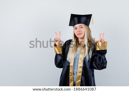  blonde girl in graduation gown and cap showing peace gestures and looking cute , front view. 