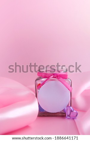 Glass jar with candy hearts inside and a pink silk ribbon on a pink background. Concept for Valentine's Day. Mockup with a place for a logo. Vertical banner. Side view. Place for text.