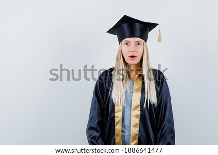  young female looking at camera in graduate uniform and looking shocked , front view. 
