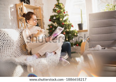 Reading magazine with her little puppy near Christmas tree. Young woman enjoying her domestic life. Home comfort, winter and holidays time concept, warm and cozy atmosphere, lifestyle, New Year.