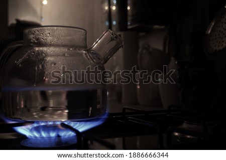 Transparent glass tea kettle on gas oven at home in the evening. Clean water heat close up background picture. Blue fire at home kitchen. Cozy winter warm evening drink, domestic cook steam